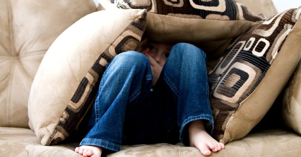 A child crouched under cushions on a couch, conveying a sense of playfulness or fear.